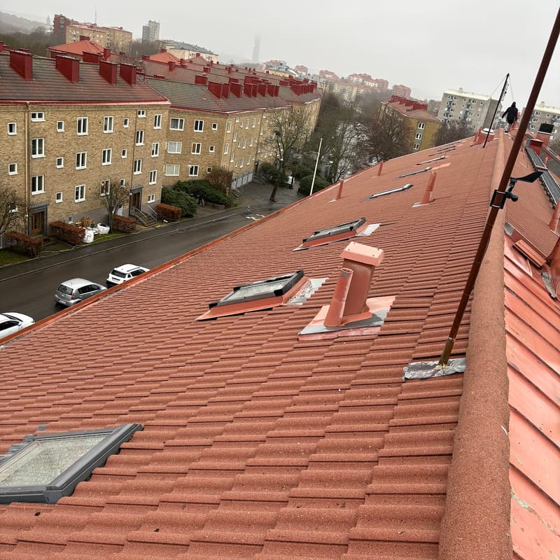 Rött tak med takpannor och takfönster på ett flerbostadshus, där en person arbetar längs nocken